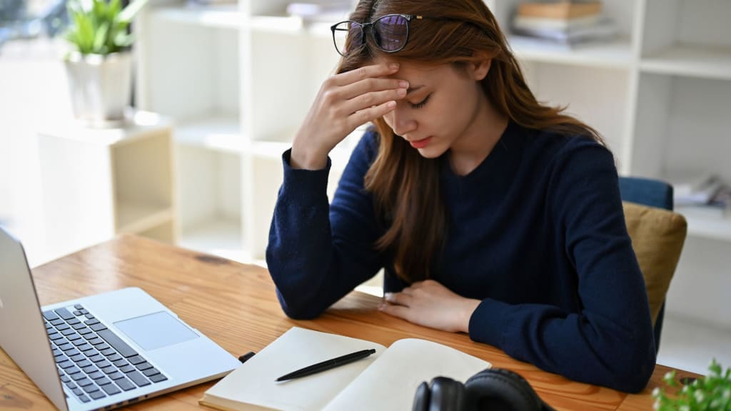 Woman sitting at a desk with a hand on her forehead, looking at an open notebook. A laptop and headphones are on the desk.
